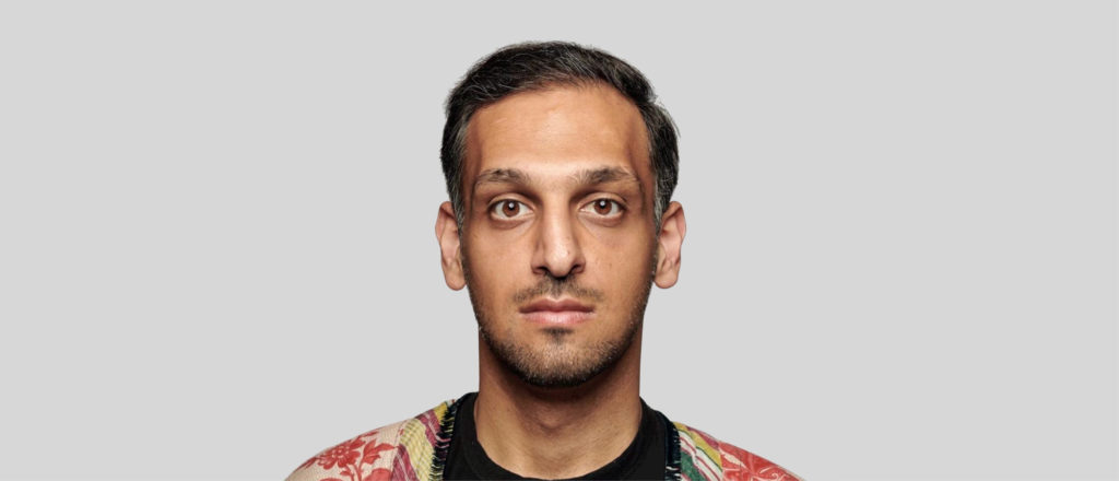 Photo portrait of Abbas Zahedi, a man with a calm serious expression looks straight at the camera, he wears a red patterned collarless shirt over a black t-shirt.